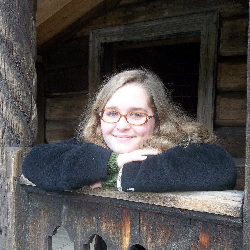 Picture. A white woman with blonde hair and glasses looking out from a Norwegian stave church.